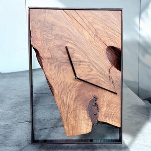 May include: A wooden clock with a black frame and black hands. The clock face is made of a natural wood slice with a unique shape and grain pattern.