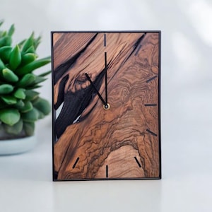 May include: A rectangular wooden clock with a black frame. The clock face features a unique wood grain pattern in shades of brown and black. Black hands and hour markers complete the design. A small succulent plant is visible in the background.