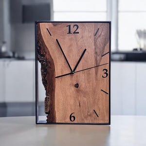 May include: Rectangular wooden clock with a natural edge and black metal frame. The clock face is light brown with black numerals at 12, 3, and 6 o'clock, and black hands. The clock is a decorative home decor item.