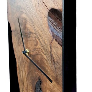 May include: A wooden wall clock with a black metal frame. The clock face is made of a natural wood slice with a unique grain pattern. The clock hands are black and the clock has a gold knob.