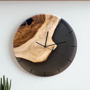 May include: A round wall clock with a natural wood and dark gray resin design. The clock face features black hands and hour markers. The wood grain is visible, adding a rustic touch. The clock is a decorative item.