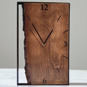 Könnte beinhalten: Eine rechteckige Holzuhr mit natürlicher Kante und schwarzen Zeigern. Das Zifferblatt zeigt die Zahlen 12, 3 und 6. Die Uhr ist in einem schwarzen Metallrahmen eingefasst, was ein modernes, minimalistisches Design ergibt.