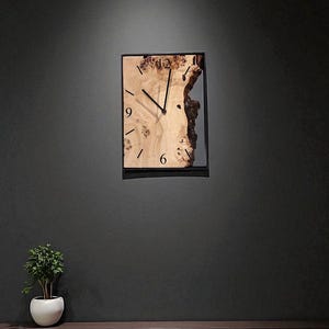 May include: A rectangular wall clock with a natural wood face and black hands. The clock's face has a light wood grain pattern with a dark edge. The clock is mounted on a dark gray wall above a wooden bench.