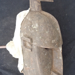 Puede incluir: Una máscara de madera con un rostro humanoide, tallada de forma estilizada. La máscara es de color marrón oscuro y tiene una textura rugosa. Está parcialmente cubierta por un paño blanco.