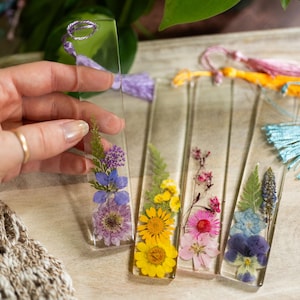 May include: Five clear rectangular bookmarks with pressed flowers and a tassel. The bookmarks feature a variety of flowers in shades of yellow, pink, blue, and purple.