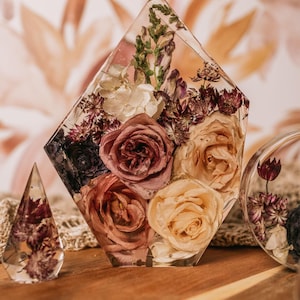 May include: A clear resin geometric display piece with preserved roses and other dried flowers in shades of pink, peach, and burgundy. A smaller, triangular resin piece and a round resin piece are also visible.