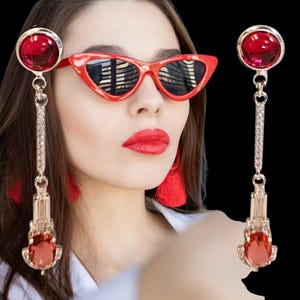 Peut inclure: Boucles d'oreilles pendantes dorées avec des pierres rouges et un motif rectangulaire. Les boucles d'oreilles sont présentées avec des lunettes de soleil rouges en forme d'œil de chat. Les boucles d'oreilles ont une grosse gemme rouge en haut et en bas.