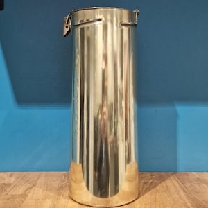 Könnte beinhalten: Ein hoher, zylindrischer, goldfarbener Metallbehälter mit einem Scharnierdeckel. Der Behälter hat eine glatte, glänzende Oberfläche und scheint aus Messing zu sein.