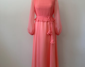 May include: A floor-length coral pink dress featuring a high neckline, vertical pleating on the bodice, and long sheer sleeves. The waist is accented with a thin tie belt that includes two small pleated tassel details hanging down the front side.