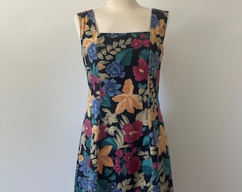 May include: A black mini dress with a square neckline and thick straps. The dress is covered in a floral pattern featuring orange, blue, and pink flowers with green leaves. The dress is displayed on a mannequin.
