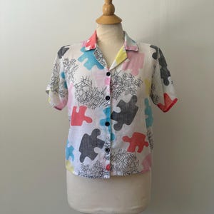 May include: A short-sleeved shirt with a white base, patterned with colourful puzzle piece shapes. The shirt has black buttons and a collar with pink, yellow, and red trim. The design also includes black scribbled lines.
