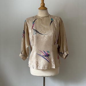 Op de afbeelding: Een beige satijnen blouse met een bloemenpatroon en driekwart mouwen. De blouse heeft een ronde halslijn en is versierd met kleurrijke paradijsvogel bloemmotieven in tinten paars, blauwgroen en zwart. De blouse wordt op een mannequin gepresenteerd.