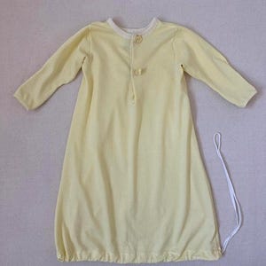 May include: A pale yellow long-sleeved gown featuring a round neckline with a light cream trim, two small decorative bows on the front placket, and a gathered hemline with a thin white drawstring cord extending from the bottom right side.