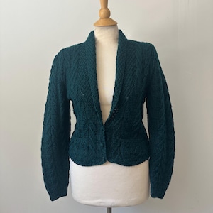 May include: A dark teal green knitted cardigan with a shawl collar and long sleeves. The cardigan is displayed on a mannequin, showcasing its textured knit pattern and tailored fit. The garment has a single button closure and two small pockets.
