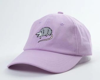 Könnte beinhalten: Eine lavendelfarbene Baseballkappe mit einem gestickten grauen und weißen Opossum auf der Vorderseite.