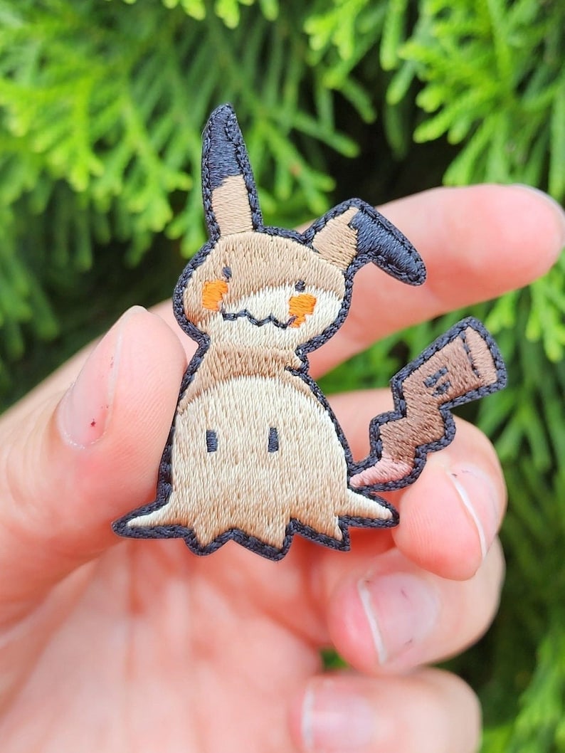 K&ouml;nnte beinhalten: Gestickter Aufn&auml;her eines braunen und schwarzen Comic-Charakters mit einem gro&szlig;en, spitzen Ohr und einem kleinen Schwanz. Der Charakter hat einen hellbraunen K&ouml;rper mit orangefarbenen Augen und einem schwarzen Umriss.