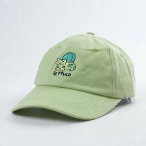 以下が含まれることがあります： つばのカーブした薄緑色の野球帽。青と緑のアクセントが付いた刺繍された漫画のキャラクターと、黒い筆記体で「lettuce」の文字が特徴です。柔らかい生地で作られた帽子です。