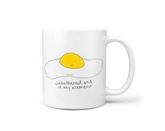 Puede incluir: Taza de cerámica blanca con un dibujo de contorno negro de un huevo frito con una yema sonriente. El texto "unbothered and in my element" está escrito debajo del huevo.