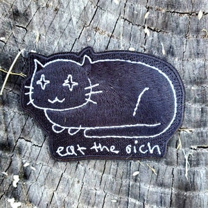 Könnte beinhalten: Schwarzer gestickter Aufnäher mit einem weißen Umriss einer Katze mit Sternenaugen und dem Text "eat the rich".