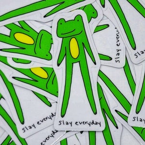 May include: A stack of green frog stickers with the text "Slay everyday" on them. The frogs have yellow bellies and are drawn in a simple cartoon style.