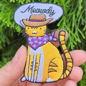 May include: An embroidered iron-on patch of a cartoon cat wearing a cowboy hat and bandana. The cat is orange and white with purple and white stars on the bandana. The text "Meowdy" is written in a speech bubble above the cat.