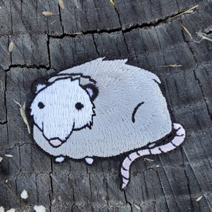 May include: A gray embroidered patch of a possum with a pink tail. The possum is facing the left side of the image. The patch is on a dark gray wooden surface.