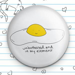 May include: White button with a black line drawing of a fried egg with a smiling yellow yolk. The text "unbothered and in my element" is written below the egg.