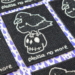 May include: Black embroidered patch with a white crow perched on a skull. The text "please no more" is stitched below the crow and skull.