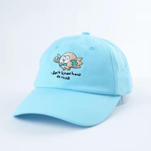 May include: Light blue baseball cap featuring an embroidered owl design with the text "I don't know how to read." The cap has a curved brim and a classic six-panel construction. A stylish and fun accessory.