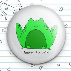 May include: A white button with a green cartoon frog and the text "born to vibe".