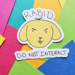 May include: A sticker featuring a yellow cartoon dog with an angry expression and the text "RABID" above its head. Below the dog is the text "DO NOT INTERACT". The sticker is on a colorful background.