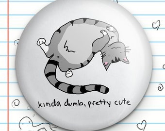Puede incluir: Un botón blanco con un gato gris de dibujos animados con rayas negras en la espalda y la cola. El gato está boca arriba con las patas en el aire. El texto "kinda dumb, pretty cute" está impreso debajo del gato.