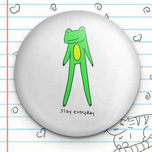 May include: A white button with a green frog illustration and the text "slay everyday".