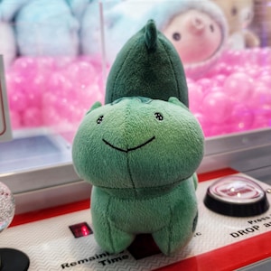 May include: Green plush toy of a cartoon character with a smiling face. The toy is sitting on a red and white surface with a button labeled "Press to Drop".