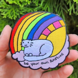 Puede incluir: Parche bordado para planchar con un gato morado sentado en una nube con un arcoíris y un sol detrás. El texto "be your own sunshine" está bordado en la nube.