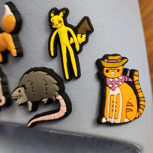 以下が含まれることがあります： 様々な漫画のキャラクターや動物が描かれた、カラフルな靴チャームのコレクション。茶色の道具を持った黄色いフィギュア、帽子とスカーフを身に着けたオレンジ色の猫、ピンクの尾を持つ灰色のオポッサムが含まれています。