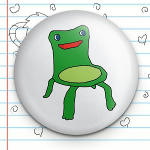 May include: A white button with a green frog-shaped chair drawn on it. The frog chair has a red mouth, blue eyes, and green legs.