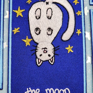 Puede incluir: Un parche bordado azul con un gato blanco colgando boca abajo contra un cielo nocturno estrellado. El texto "the moon" está bordado en blanco en la parte inferior del parche.