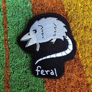 May include: A black and white embroidered patch of a cartoon opossum with the word "feral" written below it.