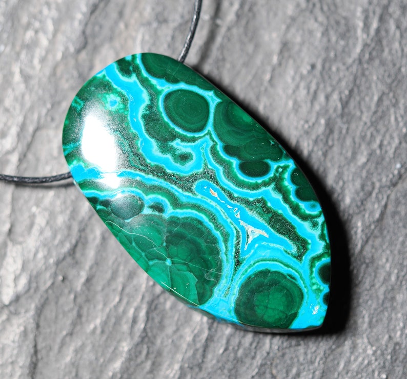 May include: A green and blue gemstone pendant with a swirling pattern. The pendant is attached to a black cord.