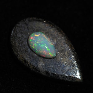 Puede incluir: Una piedra preciosa de ópalo en forma de lágrima con un brillo iridiscente verde, rojo y amarillo. El ópalo descansa sobre una roca gris oscura.