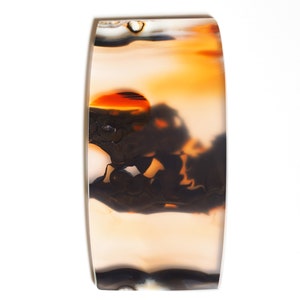 May include: A polished agate cabochon featuring a distinctive pattern of brown, white and orange. The agate has a layered appearance with a central orange area.