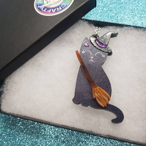 May include: A black cat with a purple hat and a broom, sitting on a white background. The cat is wearing a silver glitter hat with a purple gem. The broom is brown and has a wooden handle.