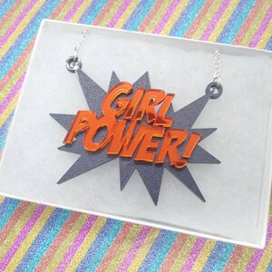 May include: A necklace with the words "GIRL POWER!" in orange, set against a gray starburst design. The necklace is displayed in a white box, with silver chain attachments.