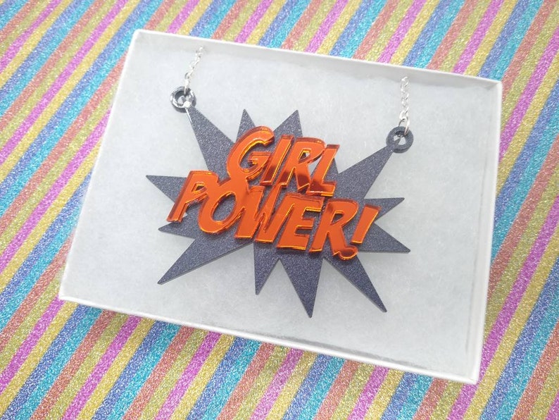 May include: A necklace with the words "GIRL POWER!" in orange, set against a gray starburst design. The necklace is displayed in a white box, with silver chain attachments.