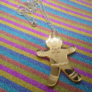 May include: A gold-colored gingerbread man pendant on a silver chain. The pendant is a cutout of a gingerbread man with a smiling face, a bow tie, and icing details.