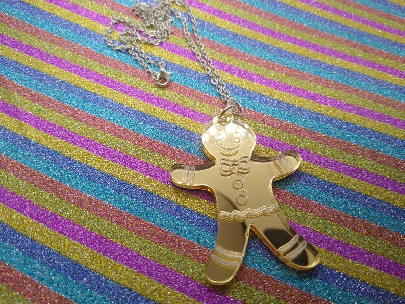 May include: A gold-colored gingerbread man pendant on a silver chain. The pendant is a cutout of a gingerbread man with a smiling face, a bow tie, and icing details.