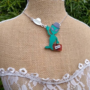 May include: A green alien figurine wearing a grey hat and holding a red suitcase with the text "AREA 51" on it. The figurine is part of a necklace with a silver chain and a silver UFO pendant.
