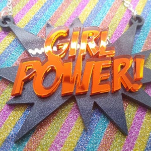 May include: A necklace pendant with the words "GIRL POWER!" in orange, reflective lettering. The text is set against a black, starburst-shaped background. The background sits on a colorful, striped surface.