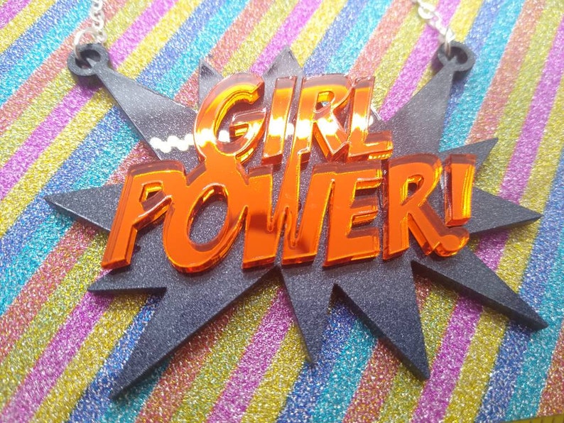 May include: A necklace pendant with the words "GIRL POWER!" in orange, reflective lettering. The text is set against a black, starburst-shaped background. The background sits on a colorful, striped surface.